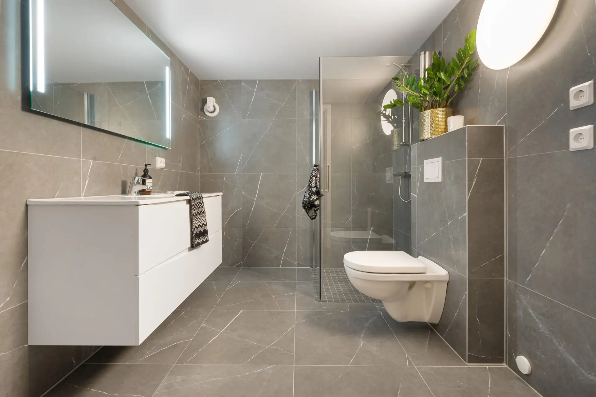 Startseite Stylish gray tiled bathroom featuring a shower, toilet, and illuminated mirror.