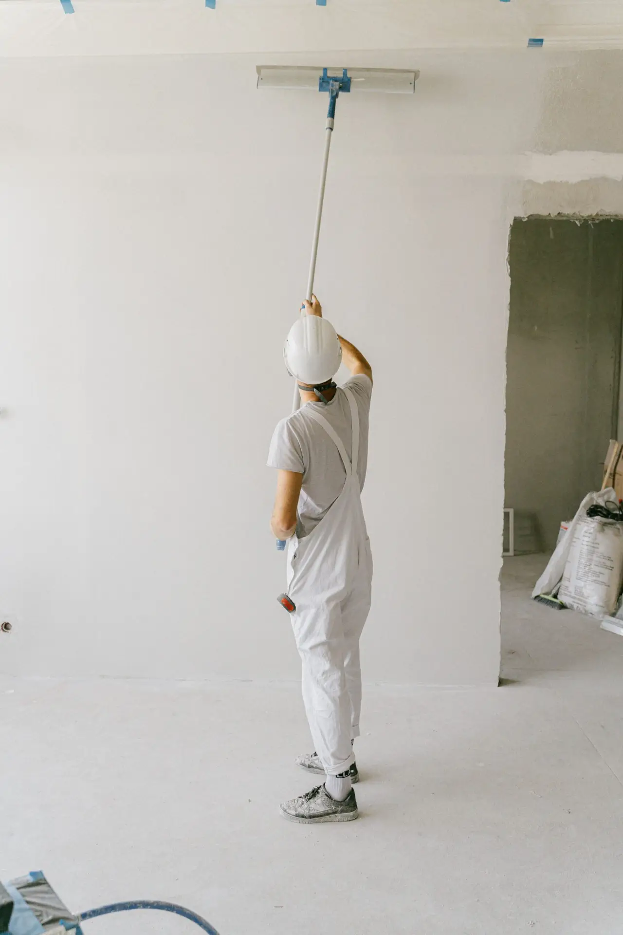 Startseite Back view of a painter in overalls and hard hat painting an interior wall with a roller.